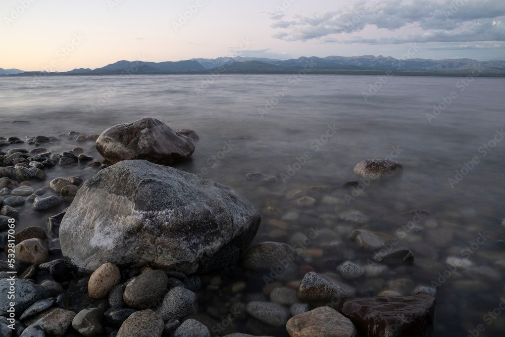 Night shot of the lake at sunset. View of the rocky shore, blurred waves and mountains in the horizon, under a colorful dusk sky. 
