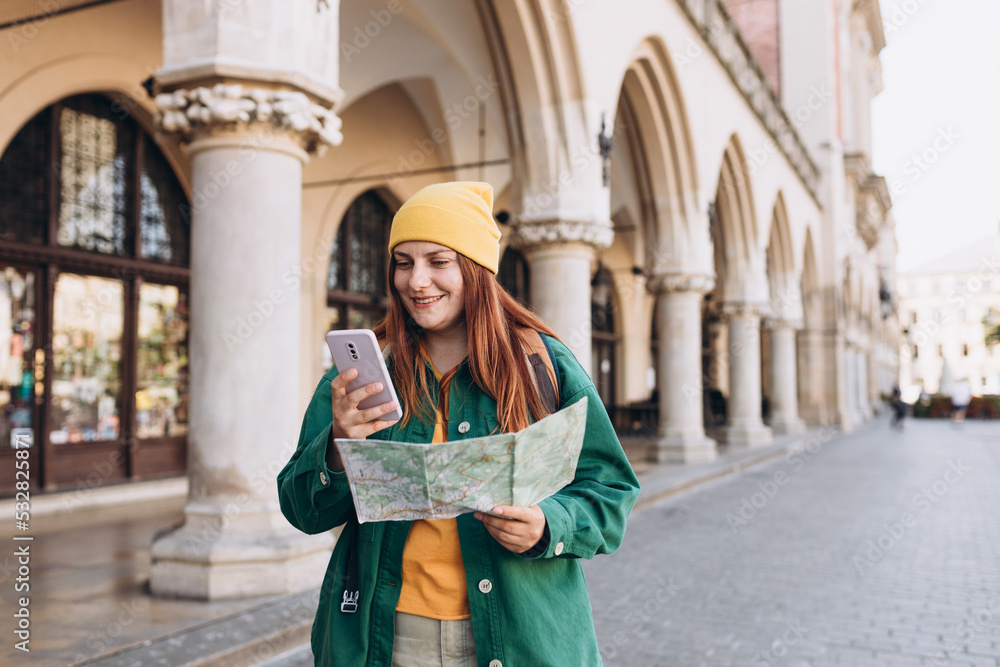 Fototapeta premium Attractive young female tourist is exploring new city. Redhead girl in hat holding a paper map and smartphone on Market Square in Krakow. Traveling Europe in autumn. Active lifestyle concept