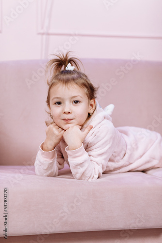 Little cute adorable Caucasian girl is lying alone on pink sofa and looking in camera in room. High quality photo