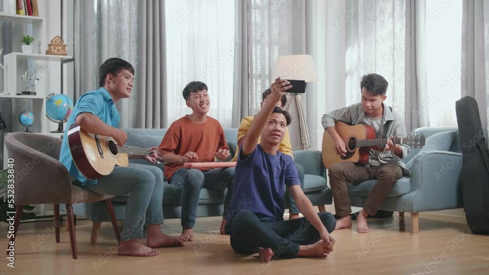 Group Of Young Asian Friends Waving Hand, Singing, And Playing Musical Instruments While Using Smartphone Lives Stream At Home
