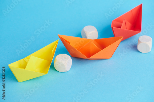 three small colorful origami boats on blue paper background circumnavigate wooden cube for example symbolism obstacles to go around, out of the way, business concept photo