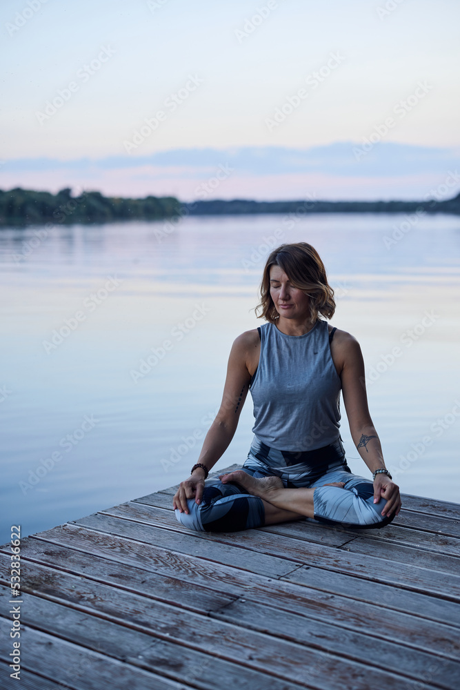A calm woman in the lotus pose sits on a dock and practices yoga breathing near the river.