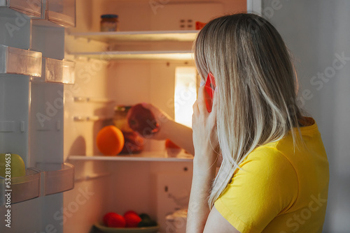Young woman opened the refrigerator and makes an application for the purchase and delivery of groceries by phone