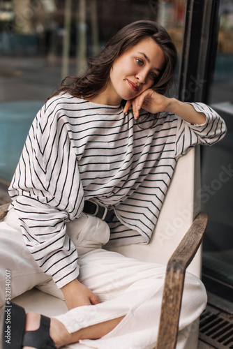 Tender young caucasian woman looks towards street sitting on armchair near city building. Brunette with wavy hair wears white sweater and pants. Relaxation concept