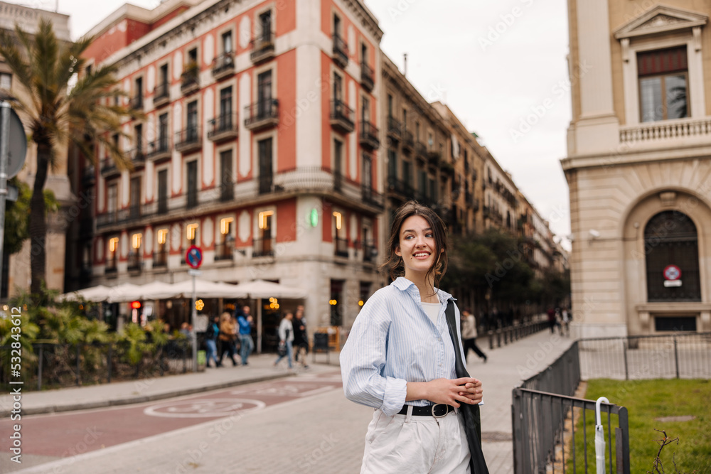 Nice caucasian young girl looks towards european city walking during day on street. Brunette wears shirt, pants and bag with her. Modern lifestyle, people and youth concept Nice caucasian young girl looks towards european city walking during day on street. Brunette wears shirt, pants and bag with her. Modern lifestyle, people and youth concept