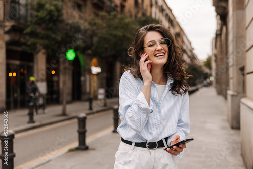 Cute young caucasian girl looks at camera, uses smartphone on street. Brunette woman with wavy hair wears casual clothes. Technology concept