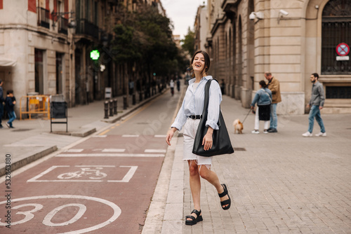In full growth, young caucasian lady spends her leisure time walking around european city on weekends. Brunette wears shirt, shorts, bag and sandals. Relaxed lifestyle, concept