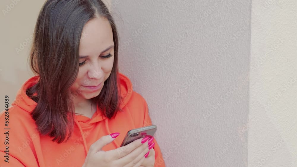 Young woman in orange hoodie browsing mobile phone, leaning against wall on street. Young woman in orange hoodie browsing mobile phone, leaning against wall on street.