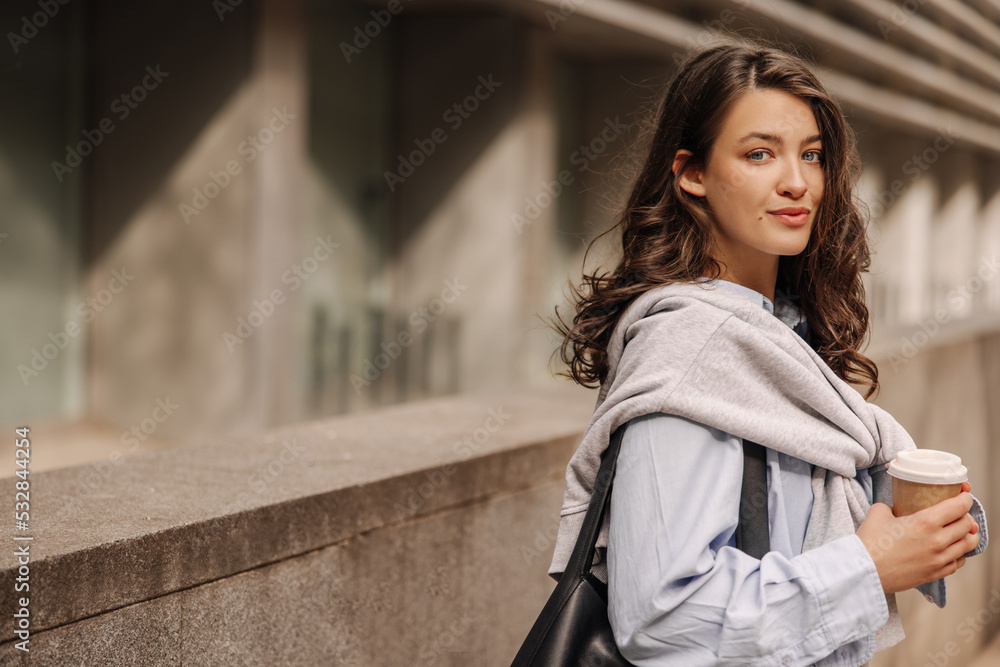 Beautiful young caucasian woman with coffee looking at camera spending time on break from work. Brunette with wavy hair wears shirt, sweatshirt and bag. Lifestyle concept Beautiful young caucasian woman with coffee looking at camera spending time on break from work. Brunette with wavy hair wears shirt, sweatshirt and bag. Lifestyle concept