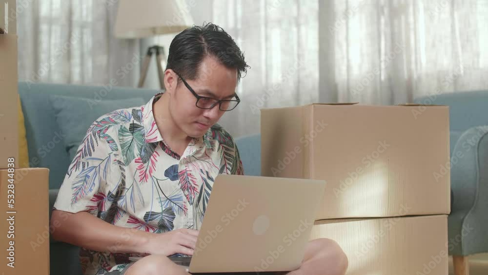 Close Up Of Asian Man With Cardboard Boxes Sitting On The Floor And Comparing The House To The Photo On A Laptop After Moving Into A New House 
