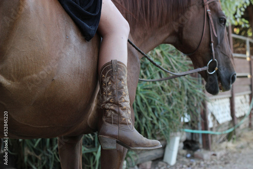 Closeup of womans leg while horseback riding in a skirt and cowboy boots on the ranch bridal path. Brown horse and brown boots bareback riding