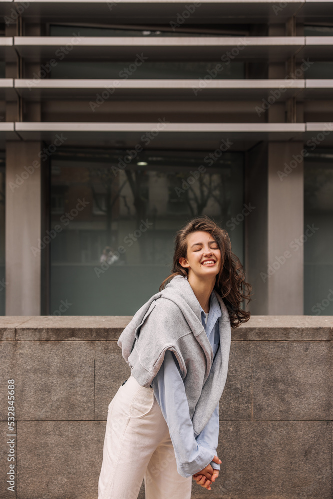 Cute young caucasian girl smiling her eyes closed spending time outdoors. Brunette with wavy hair wears shirt, trousers and sweatshirt. Holiday life concept Cute young caucasian girl smiling her eyes closed spending time outdoors. Brunette with wavy hair wears shirt, trousers and sweatshirt. Holiday life concept
