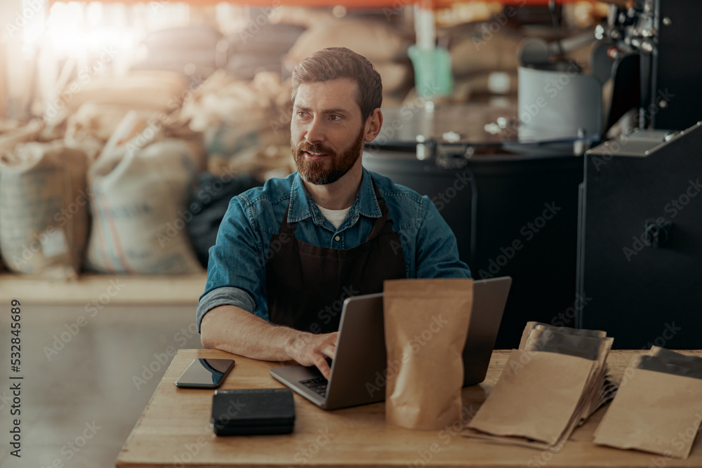 Business owner of small coffee factory working laptop on background of coffee roast machine