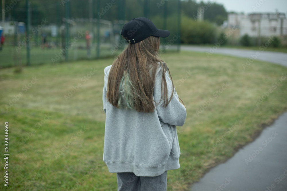 Girl in park. Teenager walks on street. Black cap and long hair. Grey jacket.
