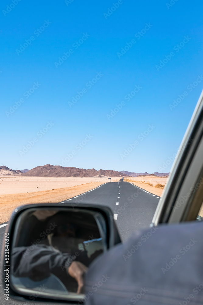Sahara Desert Road trip. Shot from the rear and outside a moving car, while the touareg driver is visible in the mirror but unrecognisable. Blurred  blue sky  and rocky mountains in Djanet, Algeria.