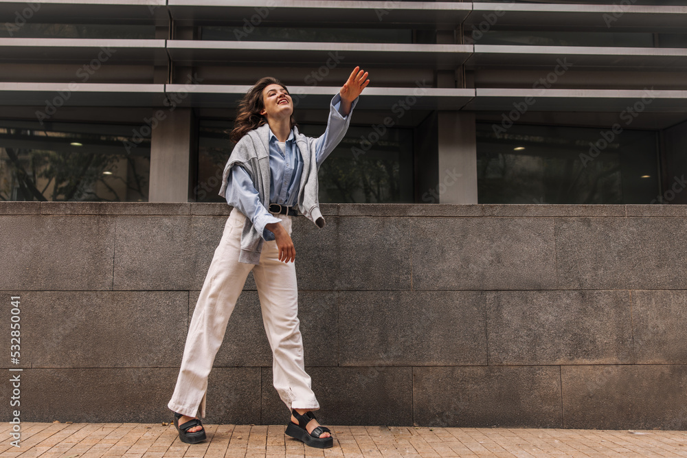 Smiling young caucasian woman in good mood spends time outdoors. Brunette with wavy hair wears shirt and trousers. Sincere emotions lifestyle concept