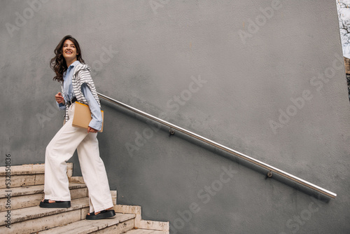 Full length positive young caucasian woman climbs stairs looking back. Brunette with wavy hair wears shirt, sweatshirt and trousers. Lifestyle concept