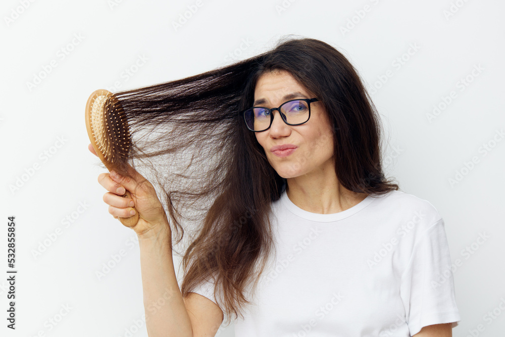 Fototapeta premium a beautiful, pleasant, sweet woman in eyeglasses and a white cotton T-shirt combs her long, well-groomed hair standing on a light background