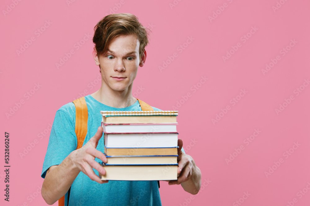 funny, red-haired student in a blue T-shirt and with an orange backpack on his back, holds a heavy stack of books in his hands and looks at the camera with surprise. Horizontal photo with empty space funny, red-haired student in a blue T-shirt and with an orange backpack on his back, holds a heavy stack of books in his hands and looks at the camera with surprise. Horizontal photo with empty space