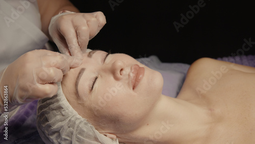 Young hypnotic face woman receiving anti-ageing facial massage in spa salon relax. Wellness body skin care face beauty treatment, rejuvenation procedure getting head face massage. Black background