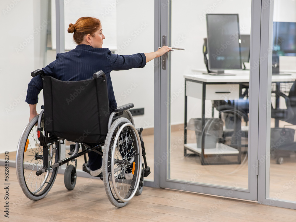Red-haired caucasian woman in a wheelchair trying to open the door in the office. 