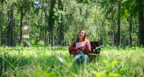 Portrait of a beautiful young asian woman reading a book and drinking coffee while sitting on a camping chair in the park