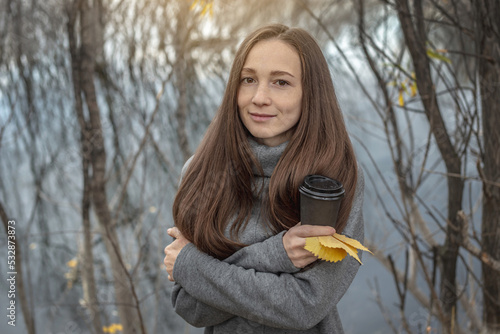 Young woman is walking through a beautiful autumn forest along the river and warming up with hot coffee