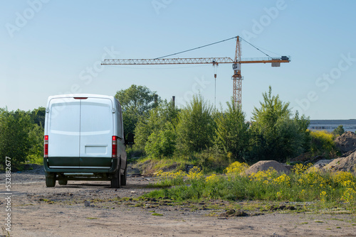 A white blank van parked on a new construction site with trees, bushes and bosk still growing, a crane erected in the background and copy space in the clear summer sky