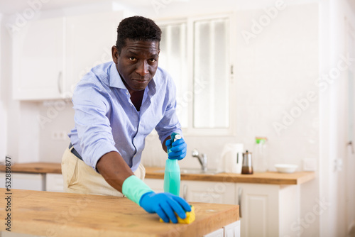Aframerican guy in gloves washes kitchen table with detergent