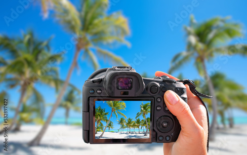 Photographer holding digital camera in hand and taking landscape picture of tropical beach with palm trees photo