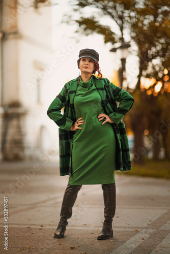 Outdoor fashion portrait of an elegant fashionable brunette woman, model in a stylish cap, green dress, posing at sunset in a European city in autumn.