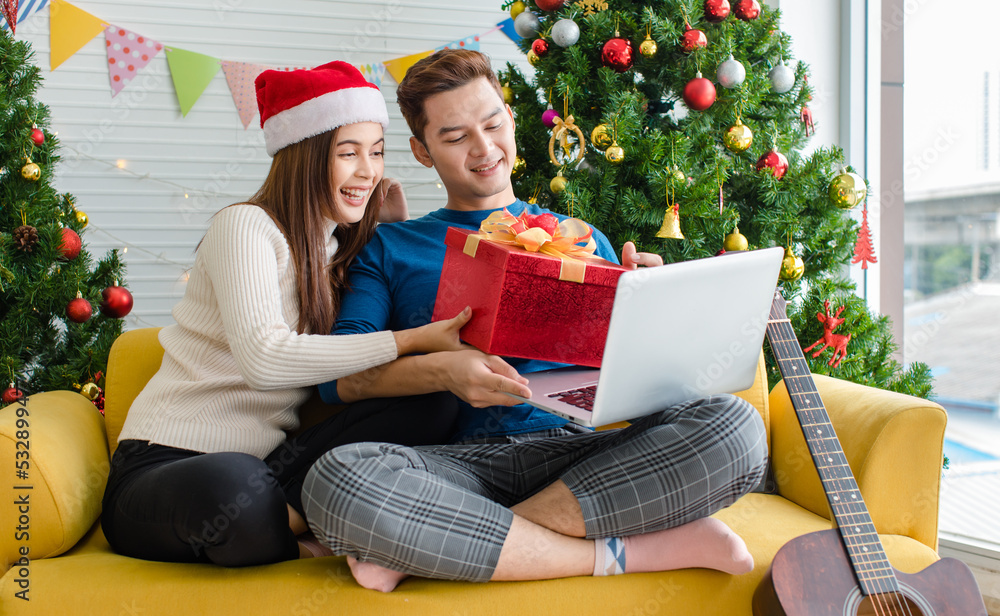 Asian happy boyfriend and girlfriend in sweater and red Santa Claus hat sitting on sofa together waving hand greeting family via laptop computer video call celebrating Christmas eve in decorated home Asian happy boyfriend and girlfriend in sweater and red Santa Claus hat sitting on sofa together waving hand greeting family via laptop computer video call celebrating Christmas eve in decorated home
