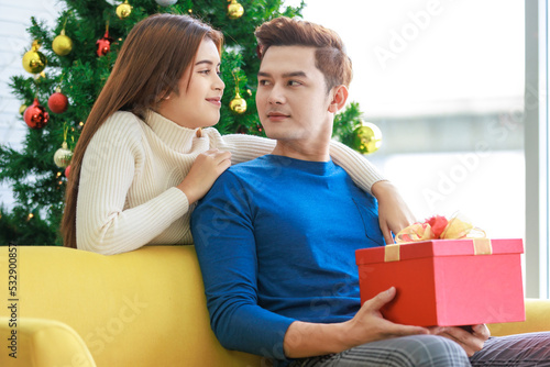Asian boyfriend sitting on cozy sofa holding opening red wrapped present gift box showing to happy girlfriend in home living room full decorated with pine trees celebrating Christmas eve festival