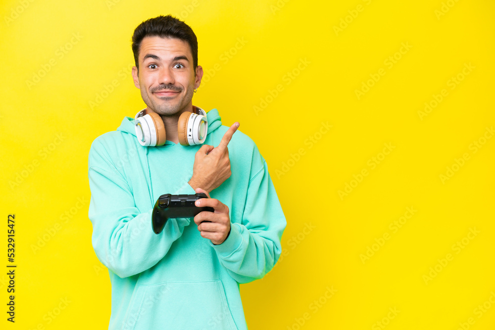 Young handsome man playing with a video game controller over isolated wall pointing to the side to present a product Young handsome man playing with a video game controller over isolated wall pointing to the side to present a product