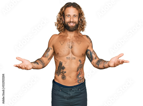 Handsome man with beard and long hair standing shirtless showing tattoos smiling cheerful offering hands giving assistance and acceptance.