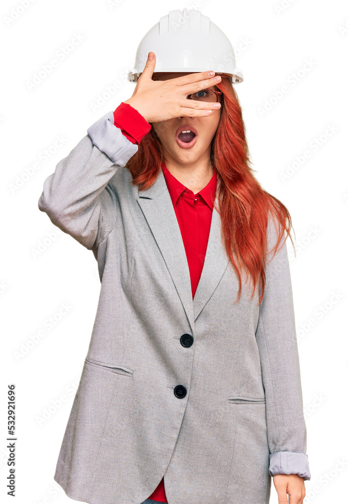 Young redhead woman wearing architect hardhat peeking in shock covering face and eyes with hand, looking through fingers with embarrassed expression. Young redhead woman wearing architect hardhat peeking in shock covering face and eyes with hand, looking through fingers with embarrassed expression.