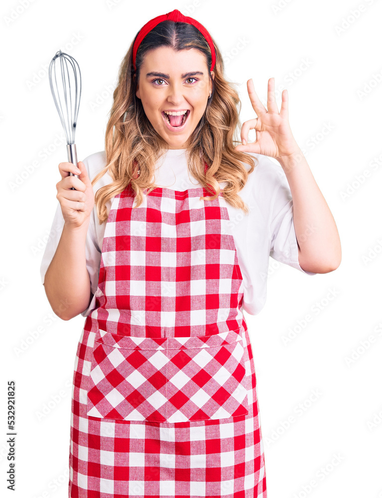 Young caucasian woman wearing apron holding whisk doing ok sign with fingers, smiling friendly gesturing excellent symbol Young caucasian woman wearing apron holding whisk doing ok sign with fingers, smiling friendly gesturing excellent symbol