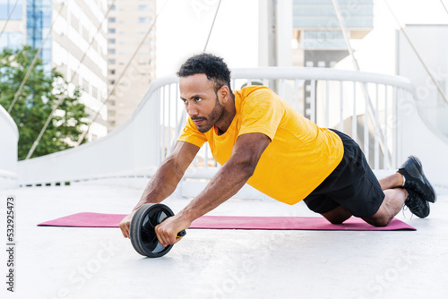 Handsome athletic african man training outdoors - Hispanic sportive man doing abs workout with ab roller wheel photo