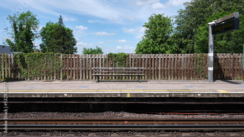 Train station platform with a wooden bench and fence. Trees and blue sky with white clouds in the background.