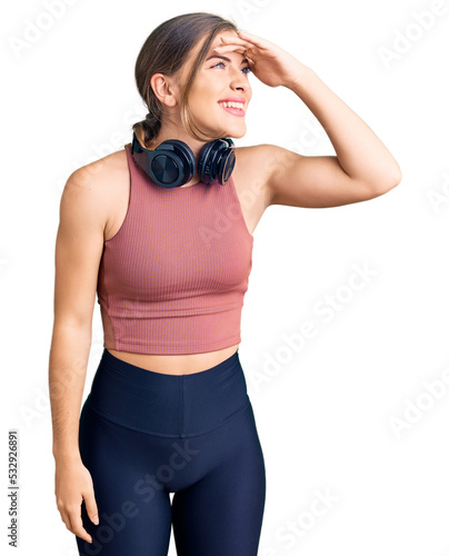 Beautiful caucasian young woman wearing gym clothes and using headphones very happy and smiling looking far away with hand over head. searching concept.