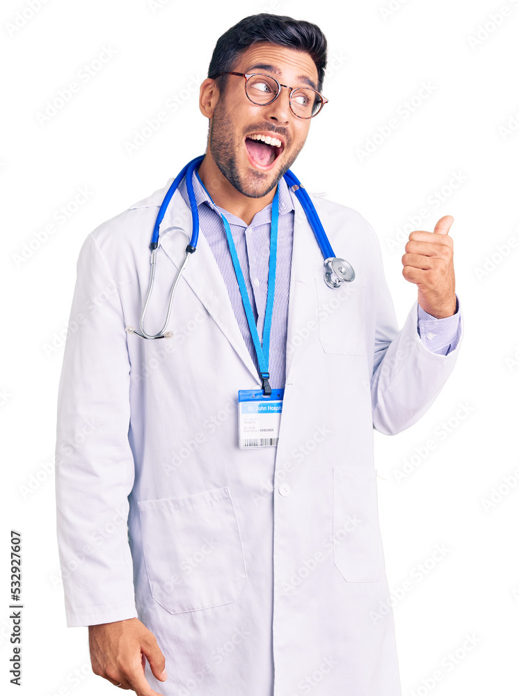 Young hispanic man wearing doctor uniform and stethoscope smiling with happy face looking and pointing to the side with thumb up.