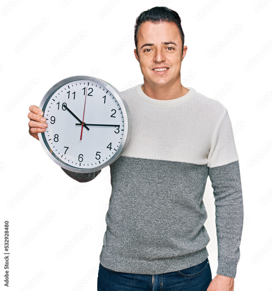 Handsome young man holding big clock looking positive and happy standing and smiling with a confident smile showing teeth Handsome young man holding big clock looking positive and happy standing and smiling with a confident smile showing teeth