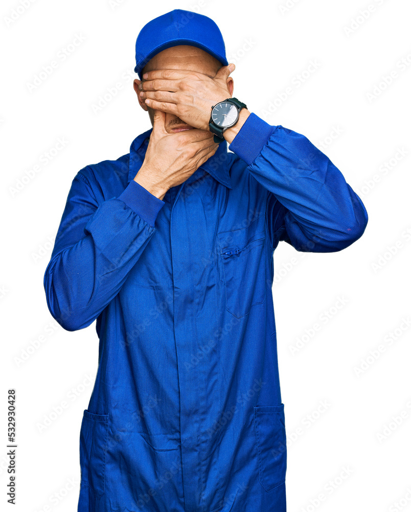Bald man with beard wearing builder jumpsuit uniform covering eyes and mouth with hands, surprised and shocked. hiding emotion Bald man with beard wearing builder jumpsuit uniform covering eyes and mouth with hands, surprised and shocked. hiding emotion