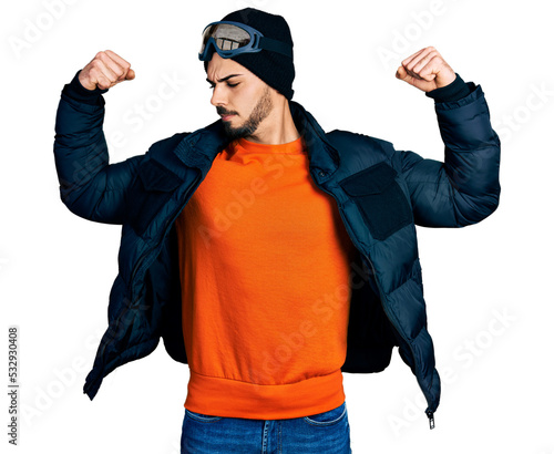 Young hispanic man with beard wearing snow wear and sky glasses showing arms muscles smiling proud. fitness concept.