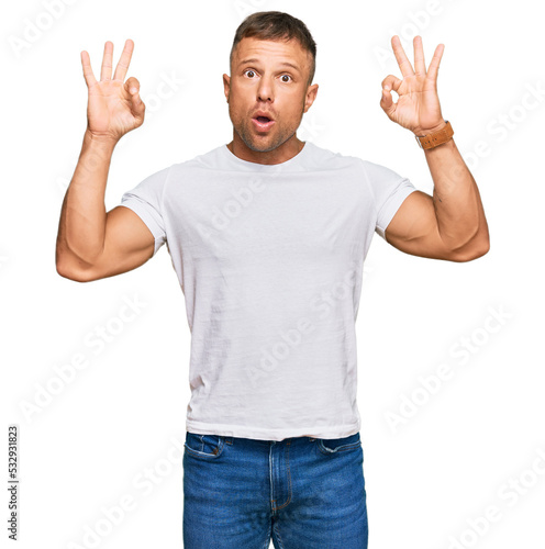Handsome muscle man wearing casual white tshirt looking surprised and shocked doing ok approval symbol with fingers. crazy expression