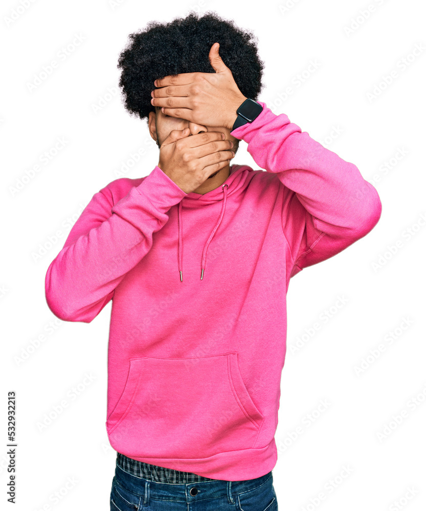Young african american man with afro hair wearing casual pink sweatshirt covering eyes and mouth with hands, surprised and shocked. hiding emotion Young african american man with afro hair wearing casual pink sweatshirt covering eyes and mouth with hands, surprised and shocked. hiding emotion