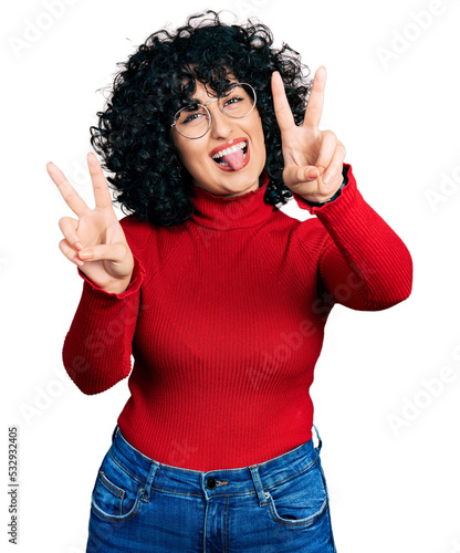 Young middle east girl wearing casual clothes and glasses smiling with tongue out showing fingers of both hands doing victory sign. number two.