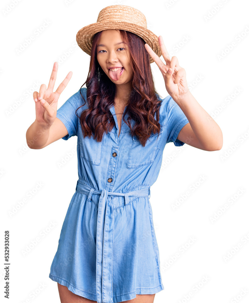 Young beautiful chinese girl wearing summer hat smiling with tongue out showing fingers of both hands doing victory sign. number two. Young beautiful chinese girl wearing summer hat smiling with tongue out showing fingers of both hands doing victory sign. number two.