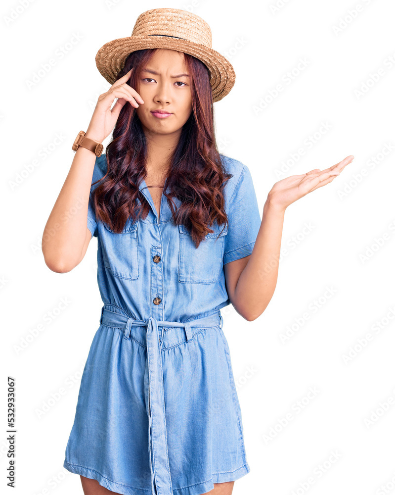 Young beautiful chinese girl wearing summer hat confused and annoyed with open palm showing copy space and pointing finger to forehead. think about it. Young beautiful chinese girl wearing summer hat confused and annoyed with open palm showing copy space and pointing finger to forehead. think about it.