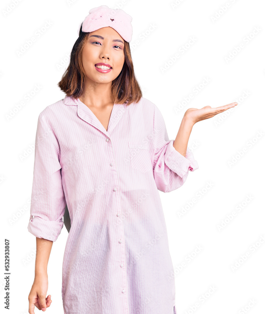 Young beautiful chinese girl wearing sleep mask and pajama smiling cheerful presenting and pointing with palm of hand looking at the camera. Young beautiful chinese girl wearing sleep mask and pajama smiling cheerful presenting and pointing with palm of hand looking at the camera.